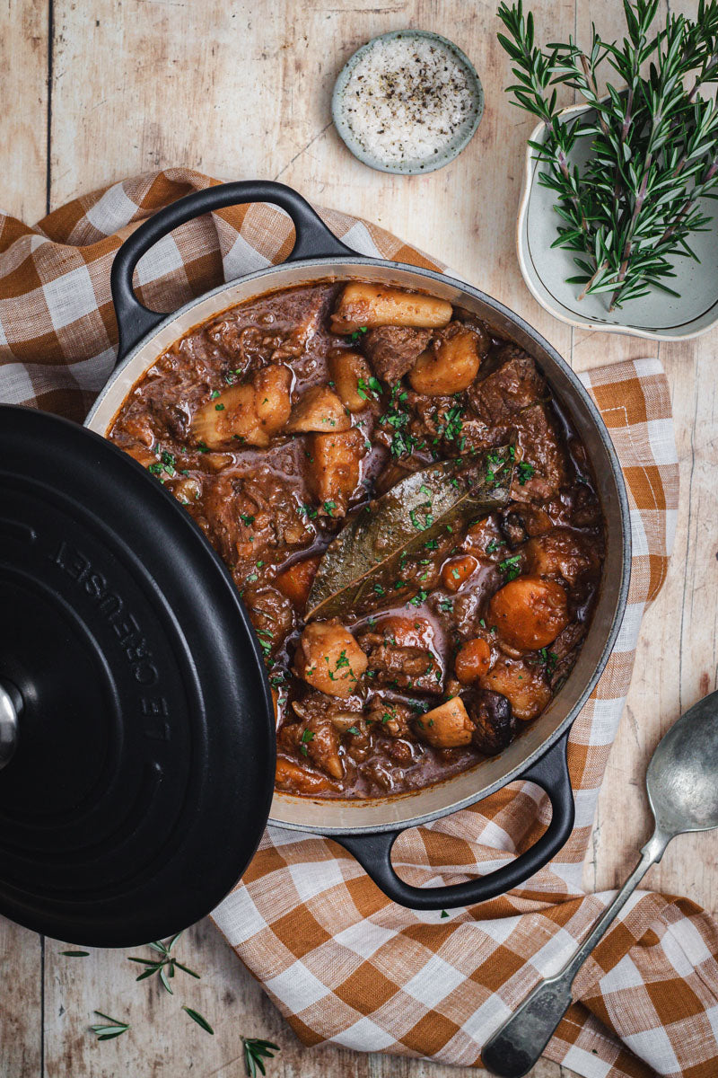 A black Le Creuset casserole with a stew baked into it. sea salt and fresh rosemary sit beside.