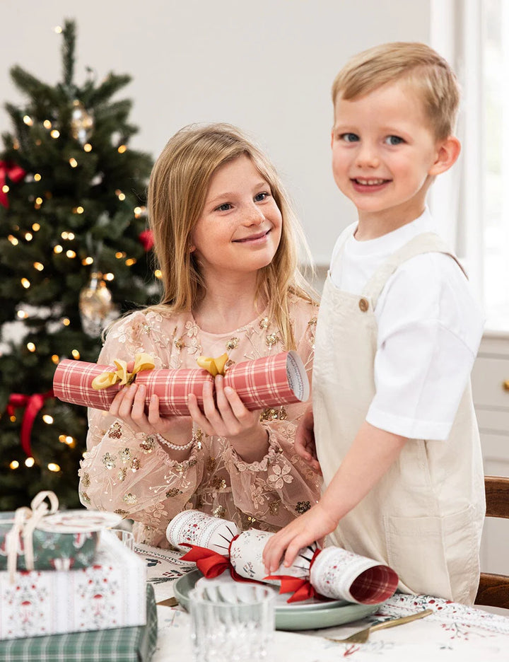 Two children holding Christmas cracker decorations in front of a decorated Christmas tree.