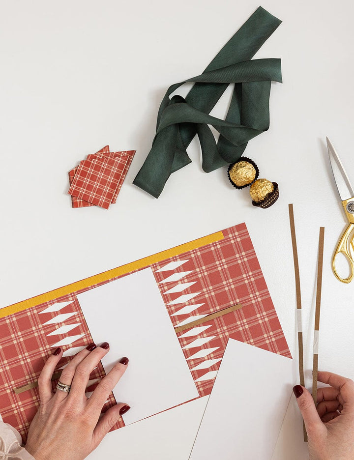 Hands working on a craft project with plaid paper, ribbons, and scissors on a white surface.