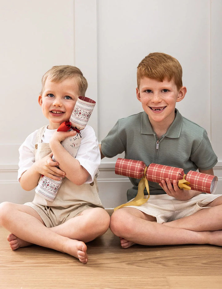 Two children sitting on a wooden floor holding Christmas crackers.