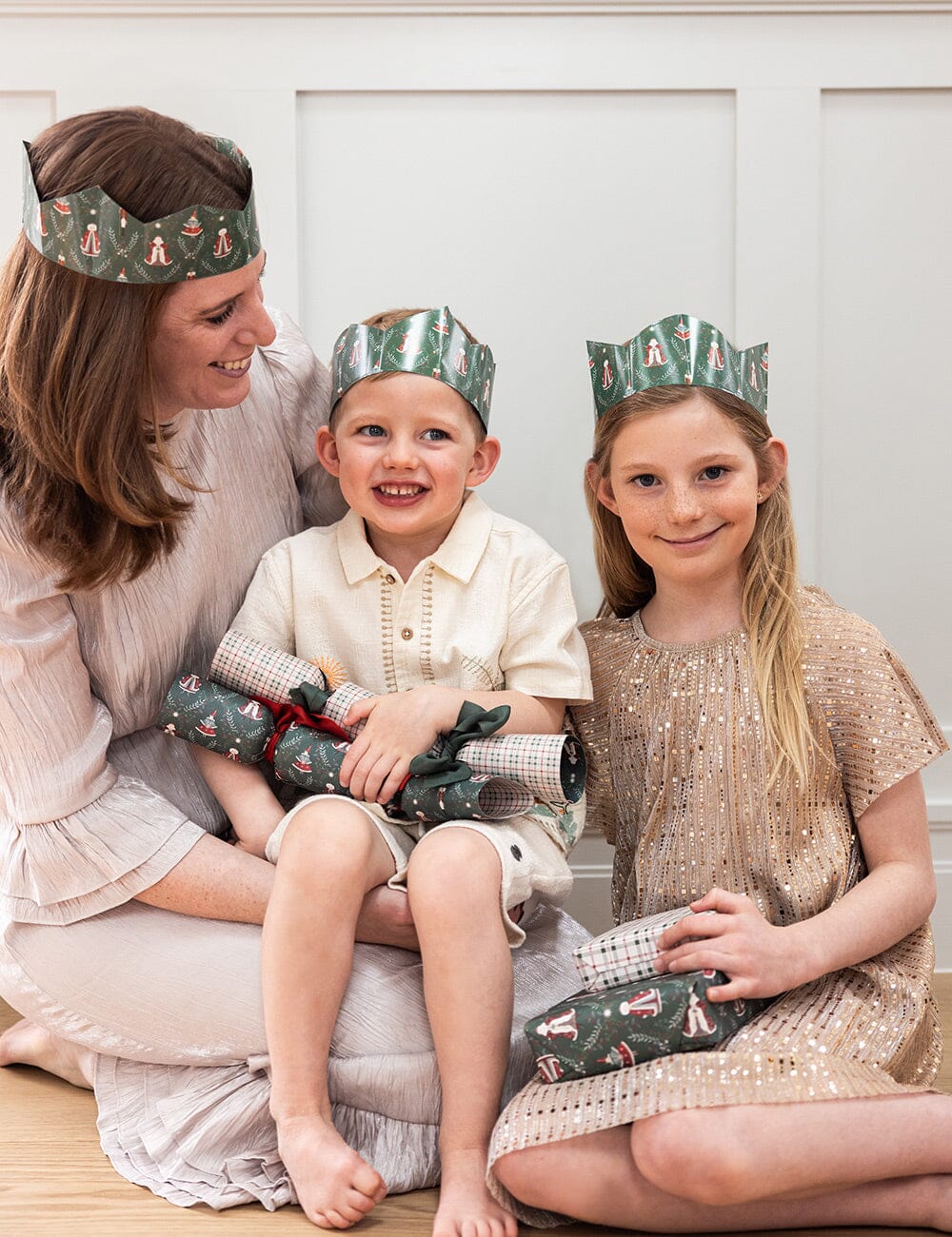 Woman and two children wearing festive crowns sitting on a white surface.