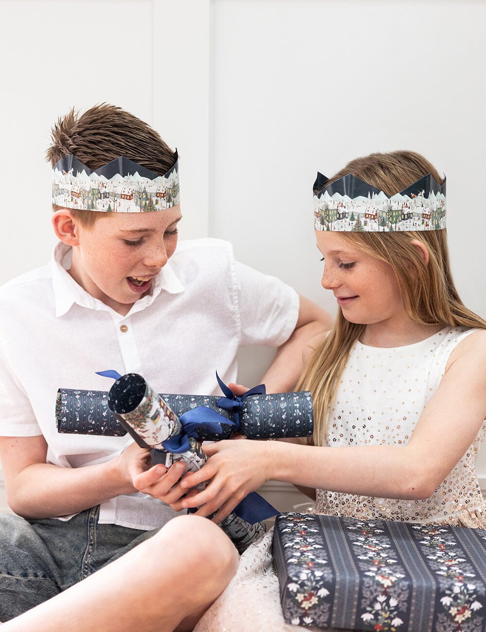Two children wearing festive headbands, opening a wrapped gift together.