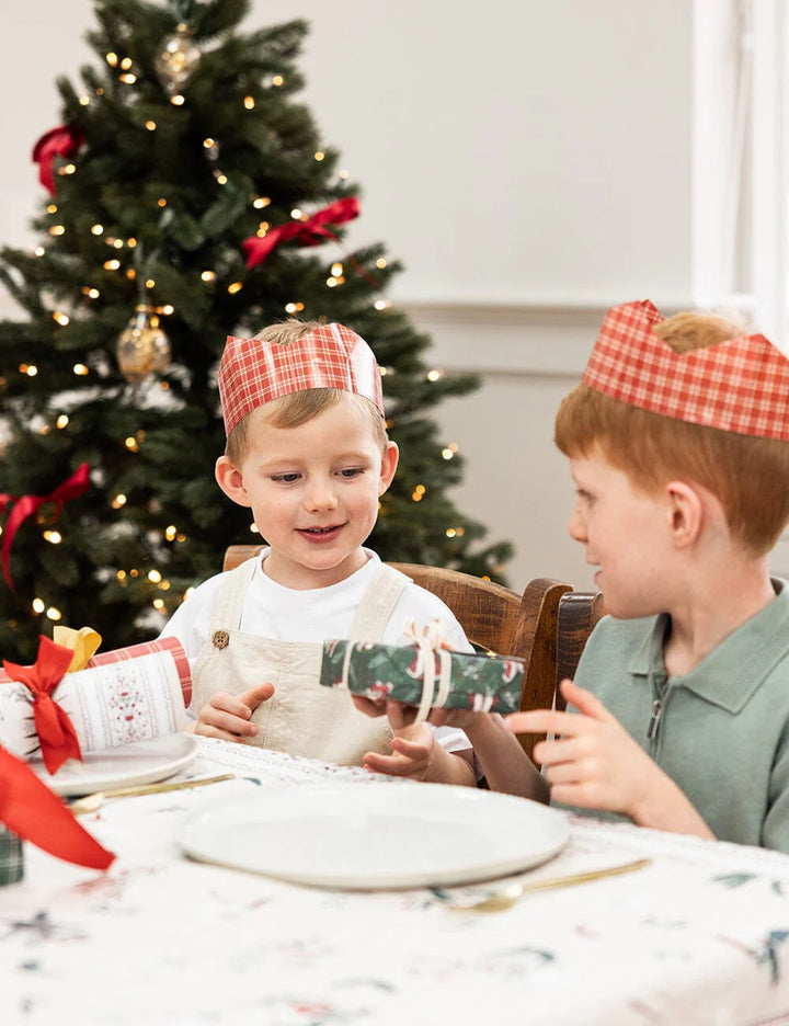 Two children at a Christmas dinner table with a decorated tree in the background.