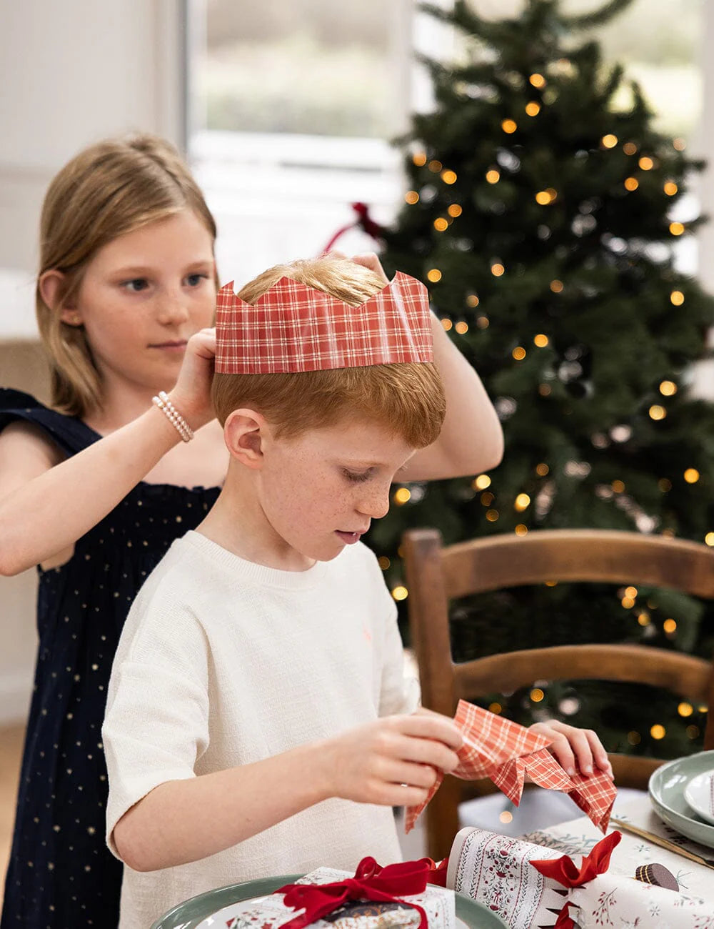 Two children in a festive setting with a decorated Christmas tree in the background.