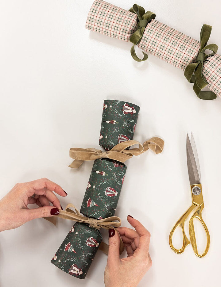 Person wrapping a Christmas cracker with green paper and gold ribbon on a white background.