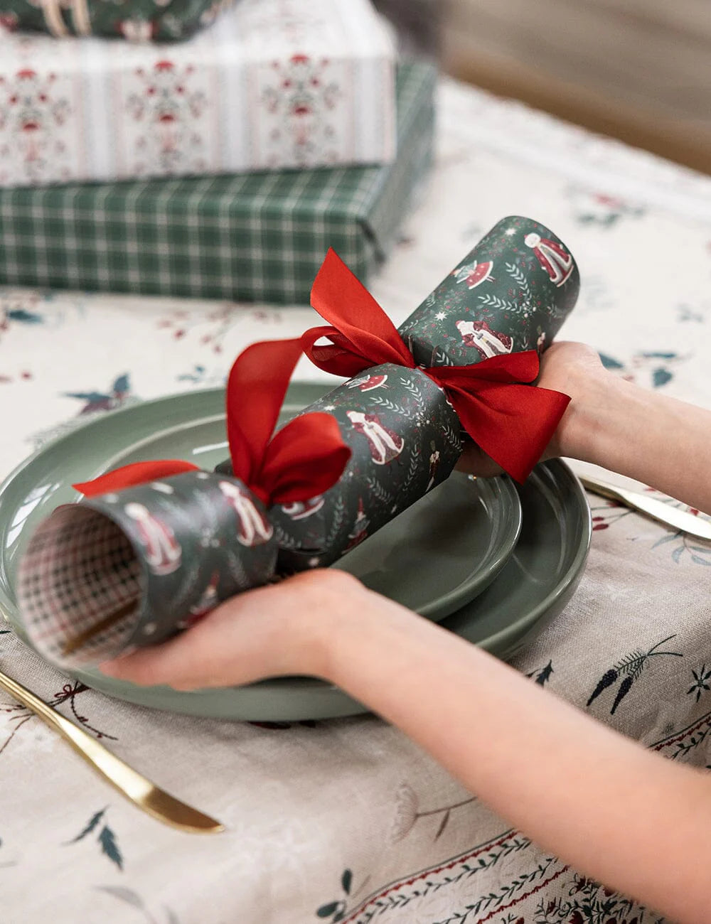 Person holding a wrapped Christmas cracker with a red ribbon on a patterned tablecloth.