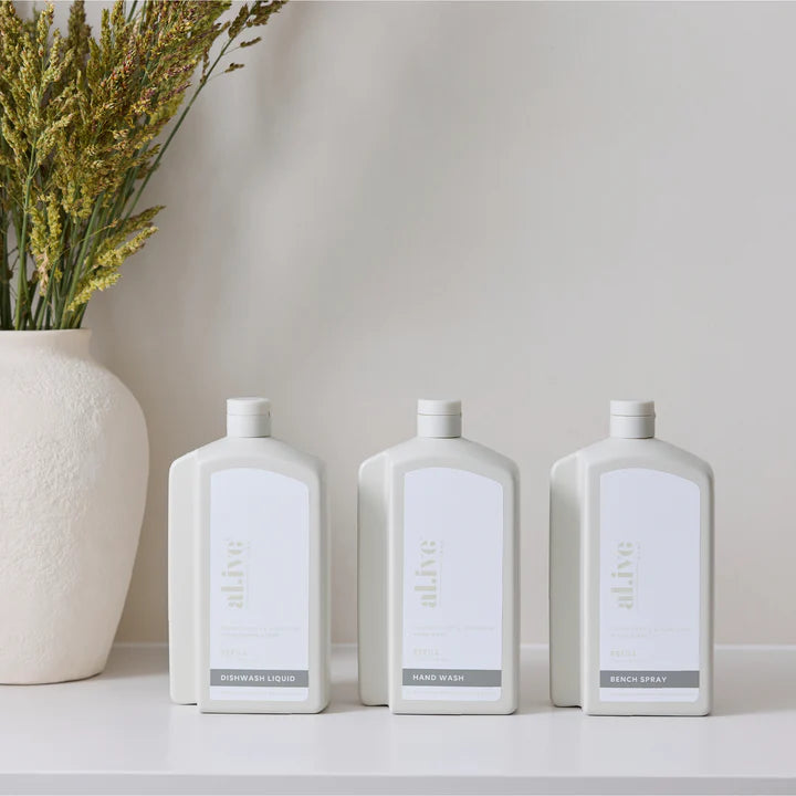 Three white bottles with labels on a light surface next to a vase with dried plants.