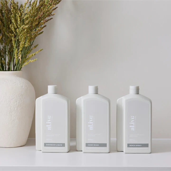 Three white bottles labeled 'Hand Wash', 'Bench Spray', and 'Dishwash Liquid' on a light surface with a vase of dried plants in the background.