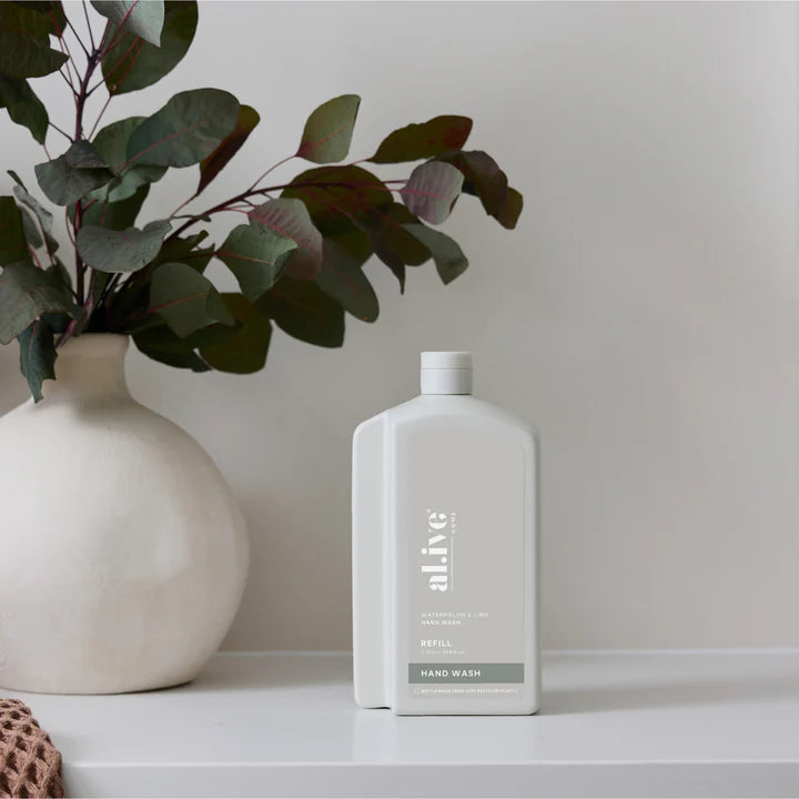 White bottle of 'alive' hand wash on a white surface with a vase and greenery in the background