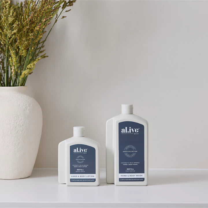 Two bottles of al.ive hand and body wash on a white surface with a vase of dried flowers in the background.
