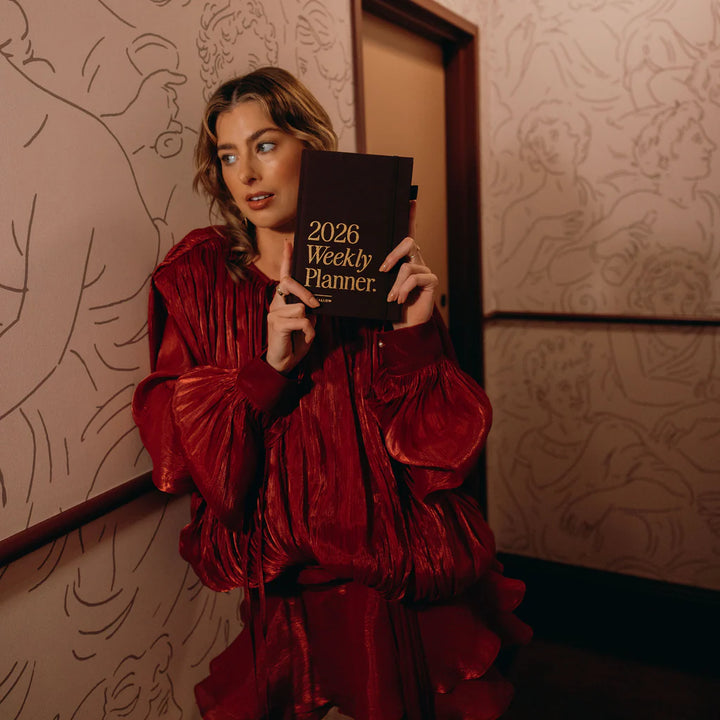 Woman in a red dress holding a 2026 weekly planner against a patterned wall.