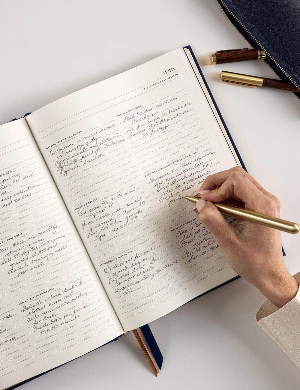 Hand writing in a notebook with a gold pen on a white surface