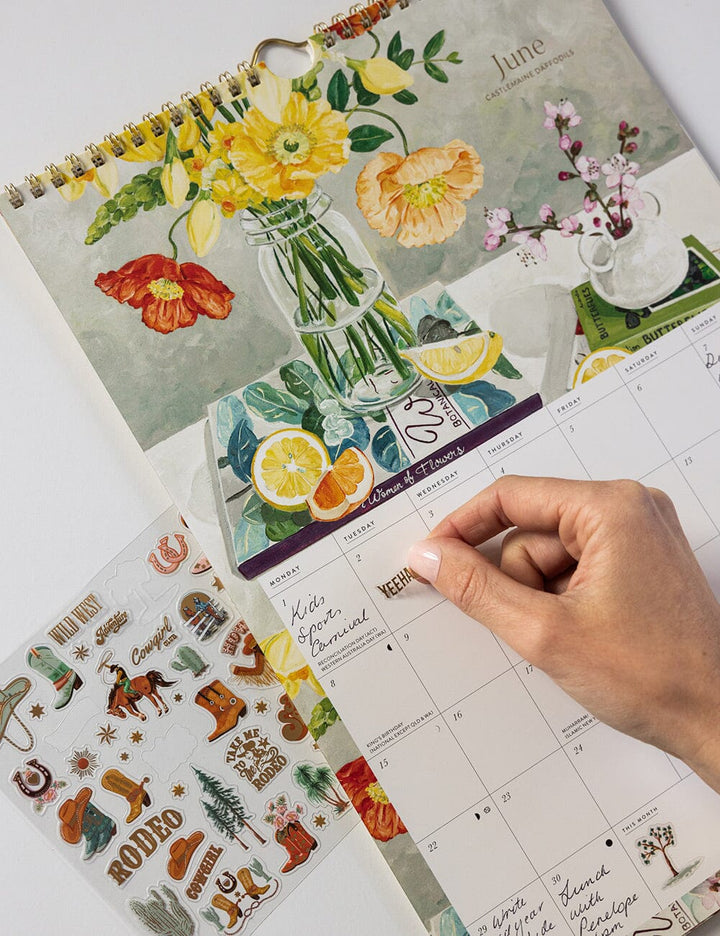 Calendar with floral designs and a hand placing a sticker on a page.