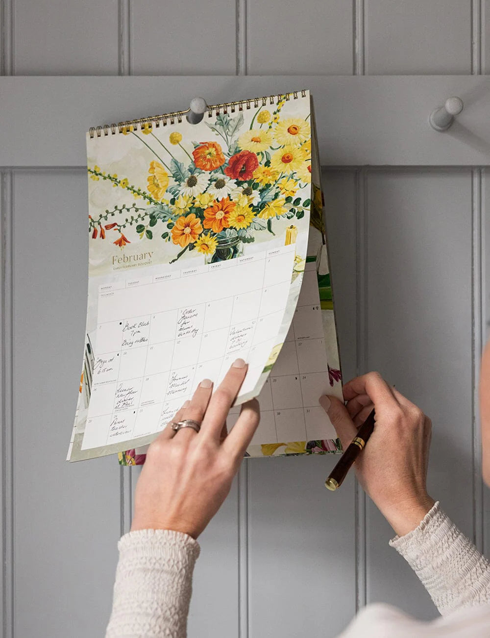 Person adjusting a floral-themed calendar on a gray wall.