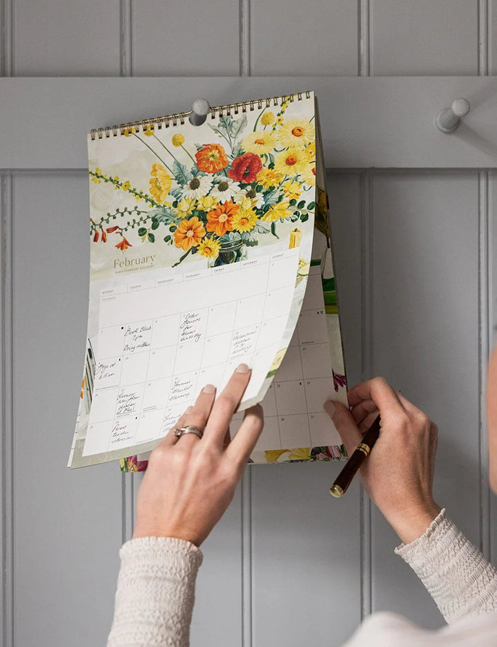 Person adjusting a floral-themed calendar on a gray wall.