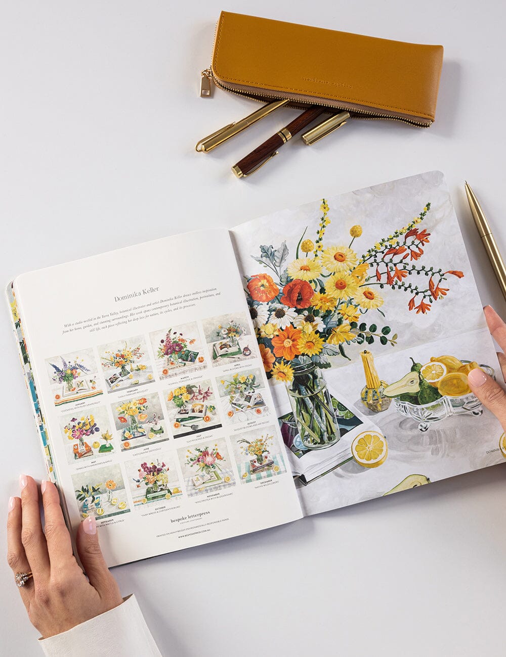 Open book with floral illustrations on a white surface with a yellow pouch and pens.