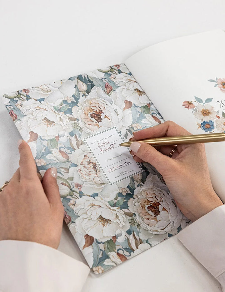 A close-up of a floral keepsake album being signed by two hands with a gold pen. The album cover features a delicate pattern of large cream-colored peonies with blue-gray and green foliage.