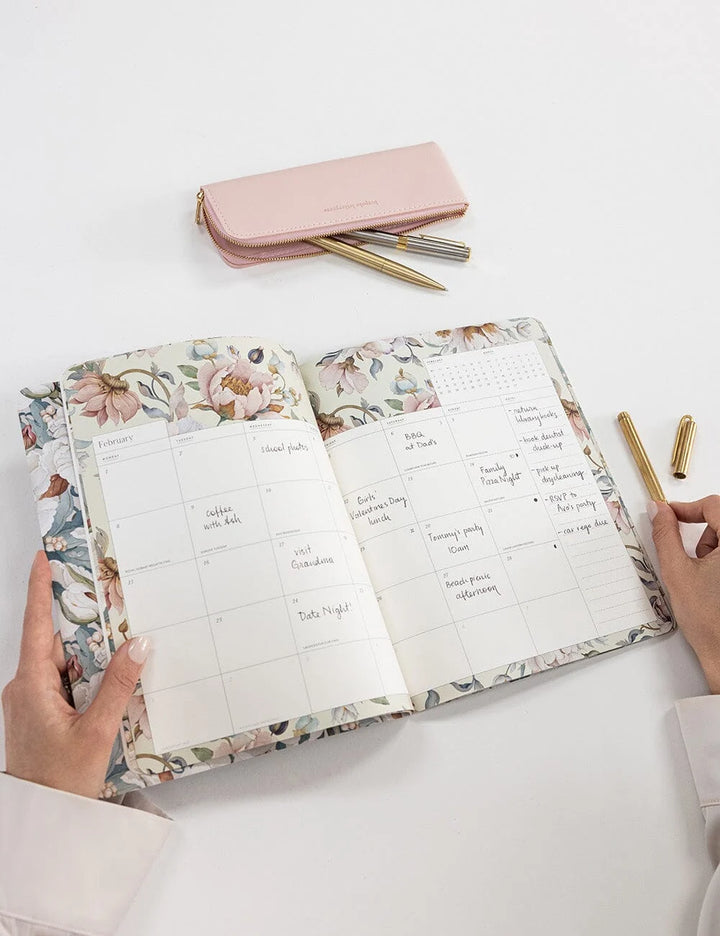 An open floral planner or agenda on a white surface, held by two hands. A pink zip pouch with gold pens and gold pen caps are placed above it.