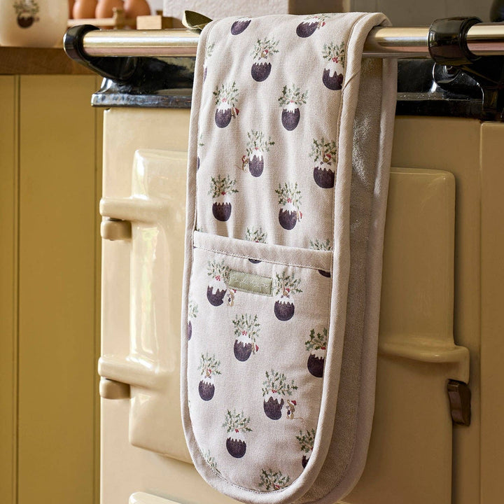 A cream-colored oven mitt with a repeated Christmas pudding pattern, hanging from a metal handle on a vintage cream-colored stove in a kitchen setting.