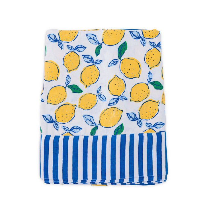 A folded linen tablecloth, featuring a yellow lemon and blue leaf pattern on a white background, finished with a blue and white striped border at the bottom.