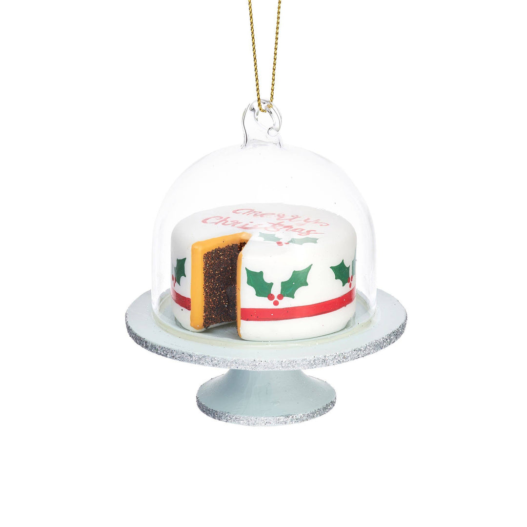 A Christmas ornament shaped like a small frosted cake under a clear glass cloche, with a slice cut out and a gold hanging string above. The cake is white with green holly motifs, a red stripe, and red lettering, displayed on a silver cake stand against a white background.