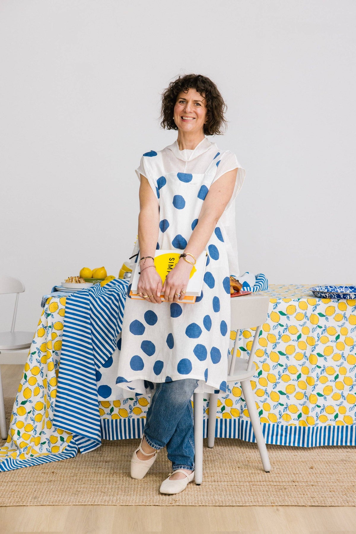Woman stands leaning against a table, wearing a white linen pinny with large blue polka dot pattern. The table is covered with a white linen tablecloth with lemon pattern, styled with blue and white striped napkin and blue bowl
