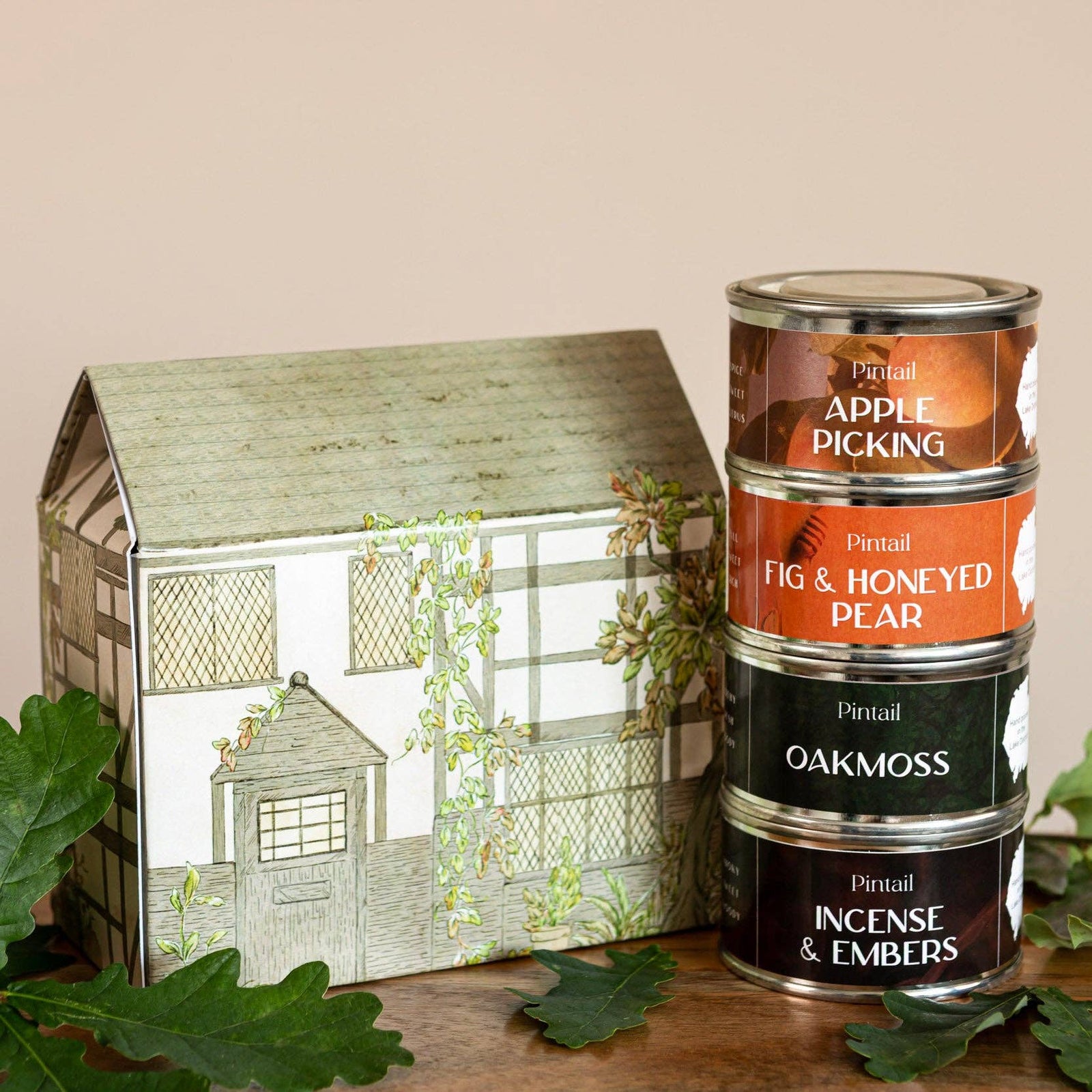 A set of four Pintail scented candles in stacked metal tins next to a decorative house-shaped gift box, styled with green oak leaves on a wooden surface against a solid beige background.