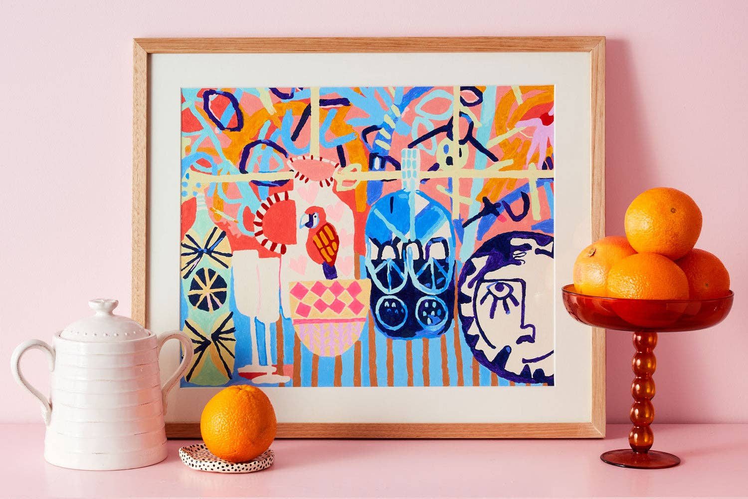 A framed abstract art print with a colorful pop-art style design featuring stylized figures and shapes in blue, pink, orange, yellow, and green, displayed on a pink surface next to a white ceramic pitcher, an orange, and a glass pedestal bowl of oranges.