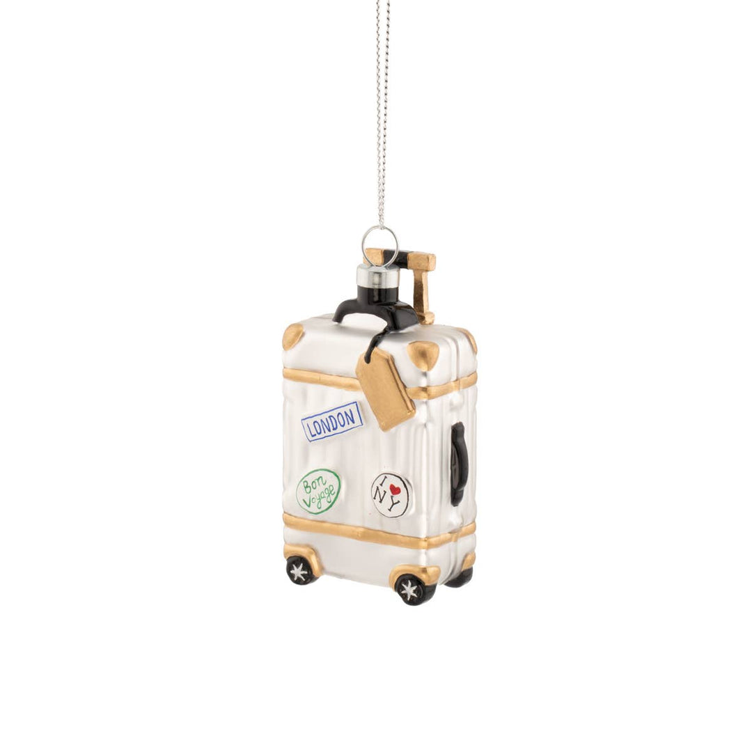 A white and gold miniature suitcase Christmas ornament with travel stickers, hanging from a silver string on a white background.