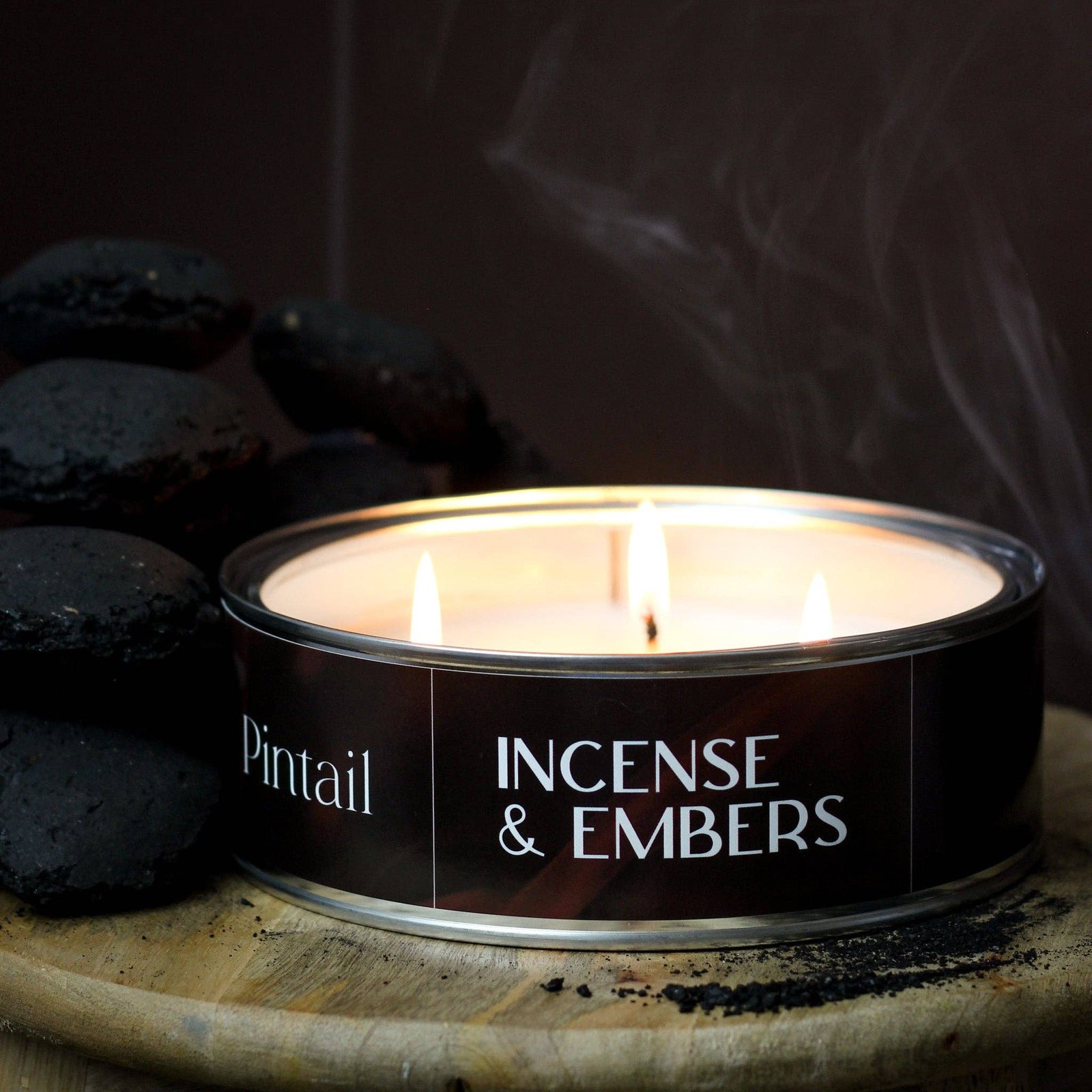 A lit candle in a round metal tin labeled 'Pintail Incense & Embers,' placed on a wooden surface with dark incense-coated stones and visible smoke in a moody setting.