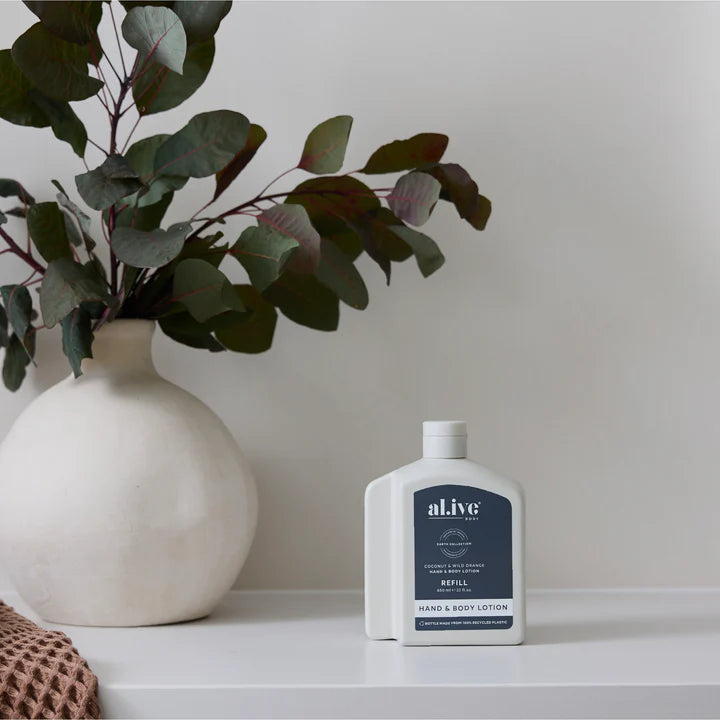 White bottle of al.ive hand and body lotion on a shelf with a vase and plant in the background.