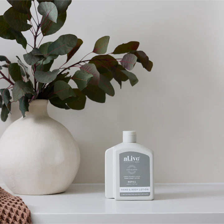 White bottle of 'alive' lotion on a surface with a vase and plant in the background