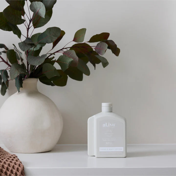White vase with green leaves and a white bottle labeled 'alive' on a light surface.