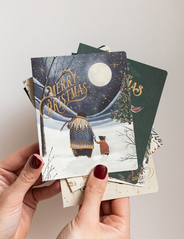 Hand holding a stack of Christmas cards with winter scenes and 'Merry Christmas' text.