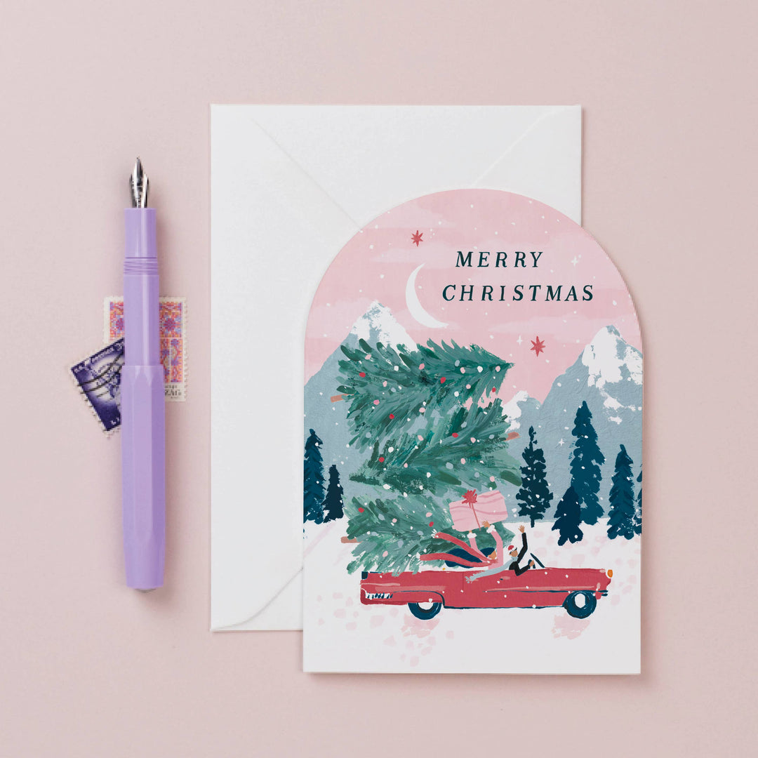 Christmas card with a red car carrying a Christmas tree on a pink background, with a pen and envelope beside it.