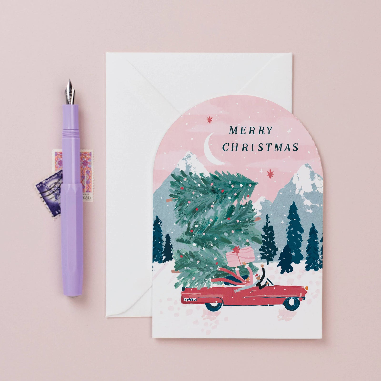 Christmas card with a red car carrying a Christmas tree on a pink background, with a pen and envelope beside it.
