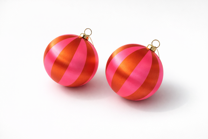 Two round Christmas ornaments with metallic pink and orange-red vertical stripes, each with a small gold hanging loop at the top, shown on a clean white background.