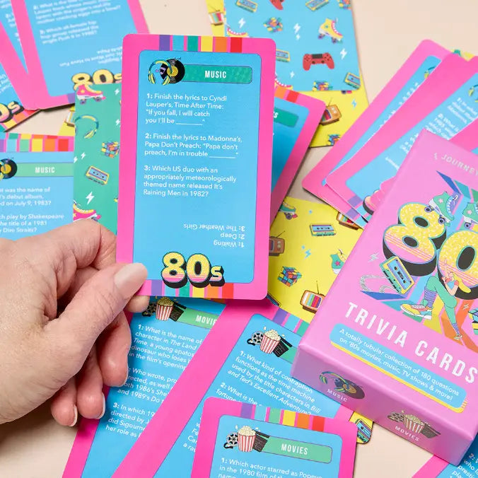 A hand holds a pink and blue trivia card from a 1980s trivia card game, with colorful cards and a pink box labeled &