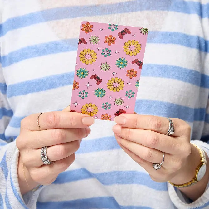 A person wearing a blue and white striped sweater holds up a pink card featuring a colorful retro pattern of flowers, stars, and small red boot motifs.
