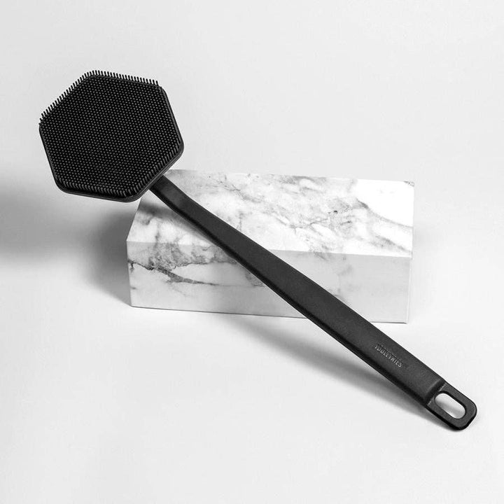 Charcoal coloured silicone back scrubber resting on a marble block, on a white background
