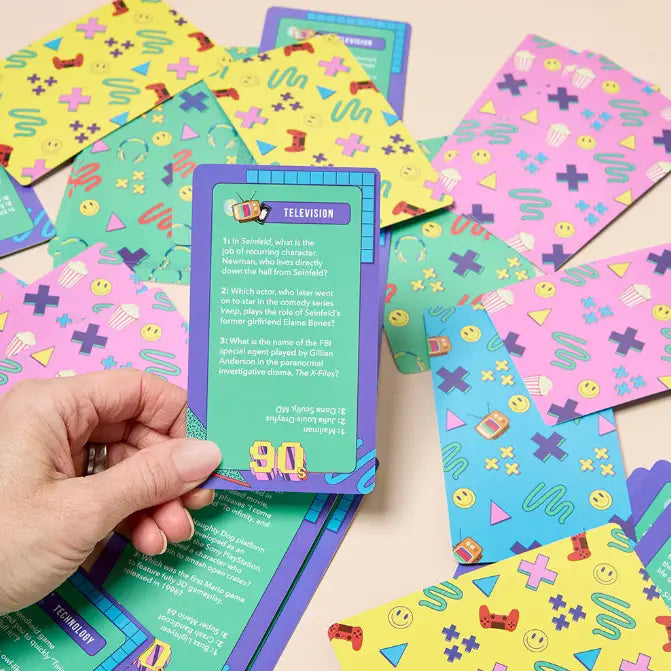A hand holds a purple and teal 'TELEVISION' trivia card from a card game, surrounded by scattered matching cards with colorful retro patterns on a light beige surface.