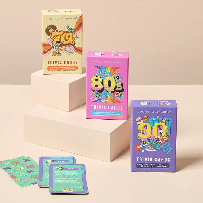 Three colorful boxed trivia card decks labeled 70s, 80s, and 90s arranged on white display blocks, with a few cards spread out in front on a warm beige studio background.
