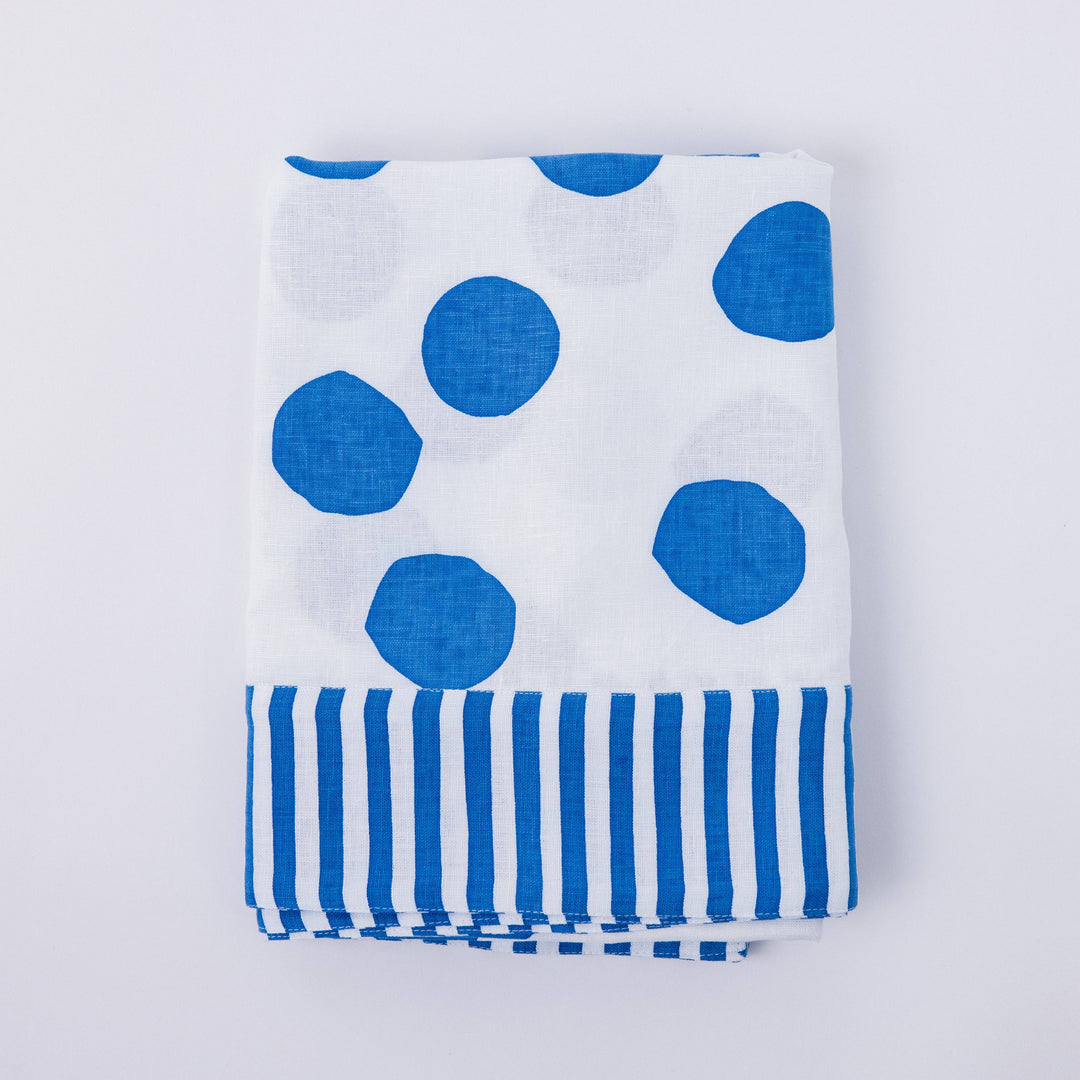 White linen tablecloth with large blue polka dots and blue striped edge pattern, folded and laid flat on a white background