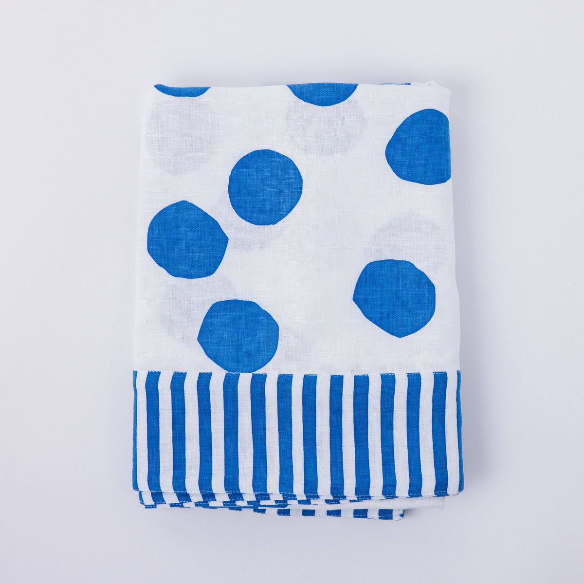 White linen tablecloth with large blue polka dots and blue striped edge pattern, folded and laid flat on a white background