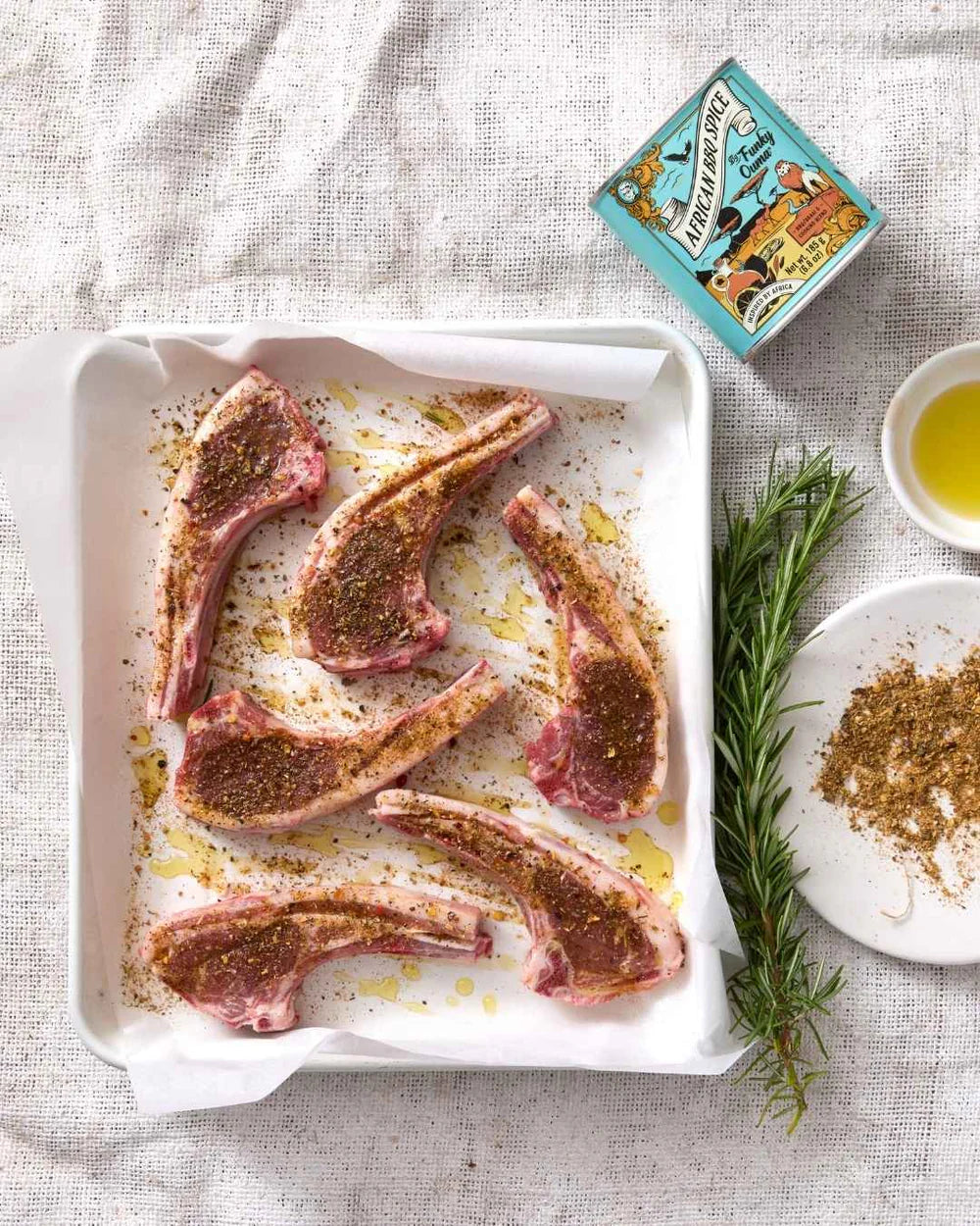 Raw lamb chops on a baking tray with seasonings and herbs on a textured surface