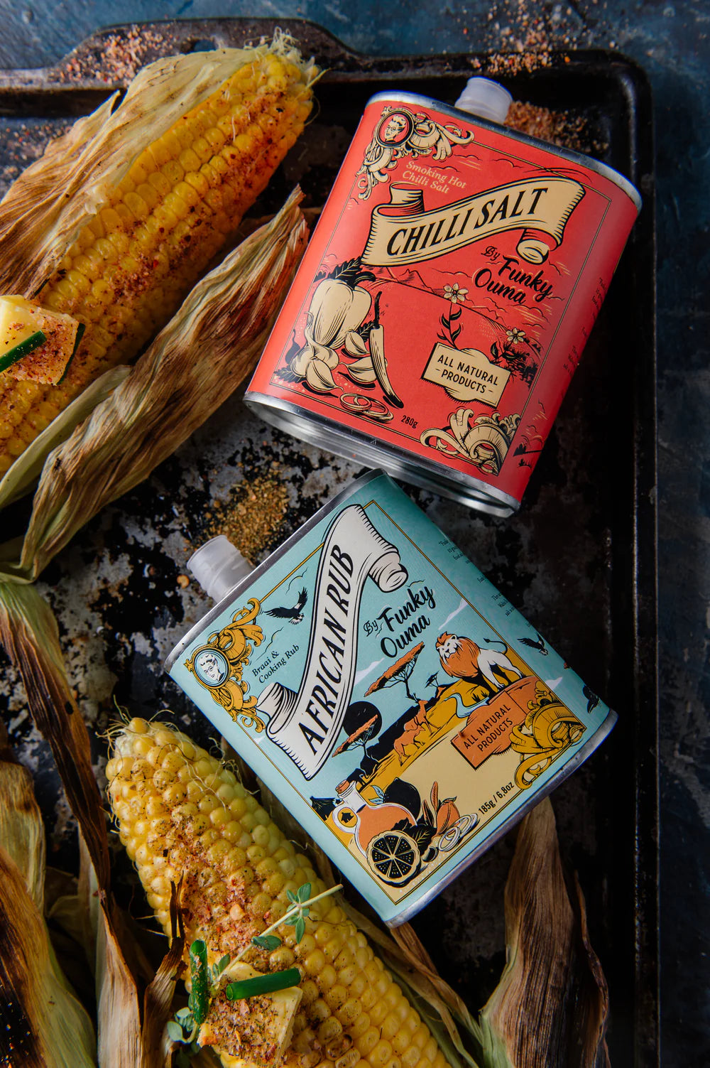 Two vintage-style tins labeled 'Chilli Salt' and 'African Rub' on a tray with corn on the cob.