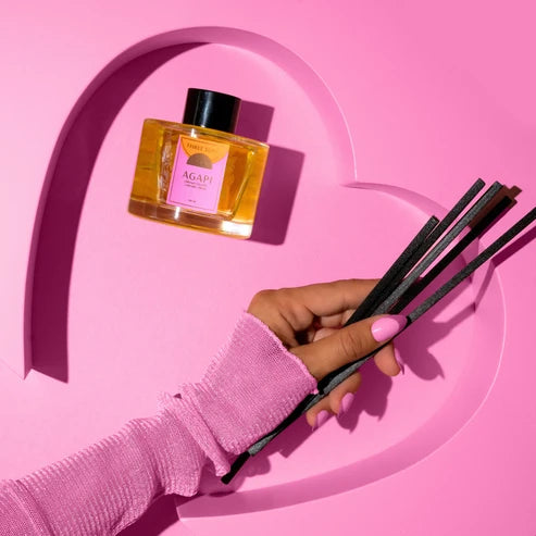 A hand wearing a pink long-sleeve top holds several dark reeds next to a clear glass bottle of AGAPI reed diffuser oil with a pink label, set against a sculpted pink background.