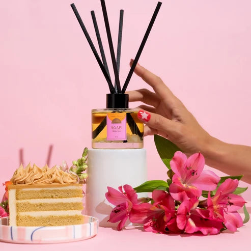 A reed diffuser bottle with black reeds and a pink label sits on a white pedestal, with a hand reaching toward it. A slice of layered cake and pink flowers are styled around it against a solid pink background.