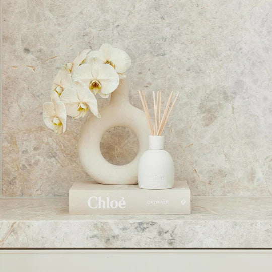 Decorative setup with a vase of flowers, diffuser, and book titled 'Chloe' on a marble surface.