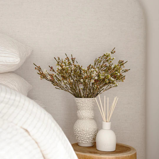 White vase with dried flowers and diffuser on a wooden table against a neutral wall.